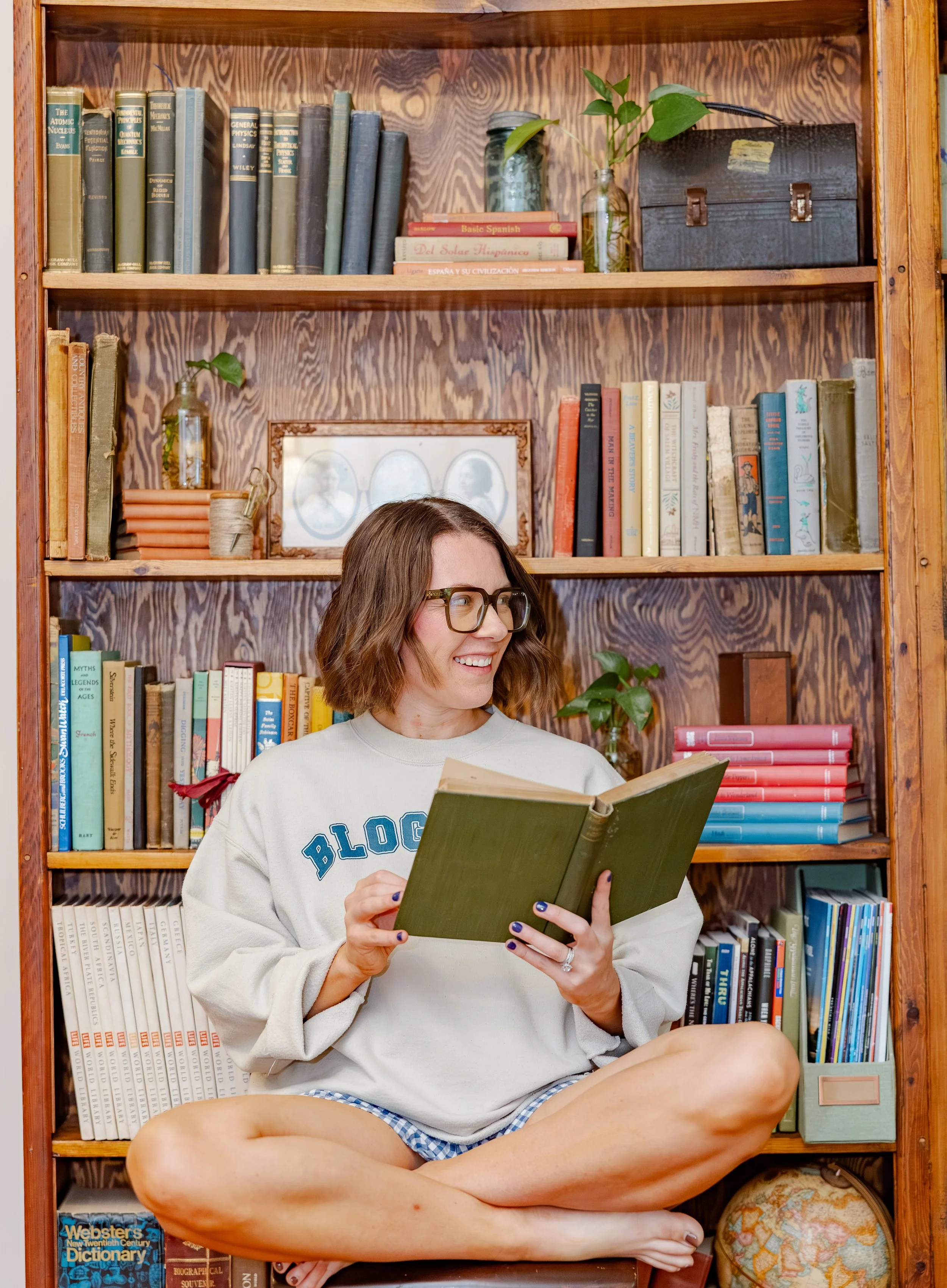 Natty Writes, business blog writer, sitting cross-legged and smiling while reading a book in front of a wooden bookshelf filled with various books and decorative items.
