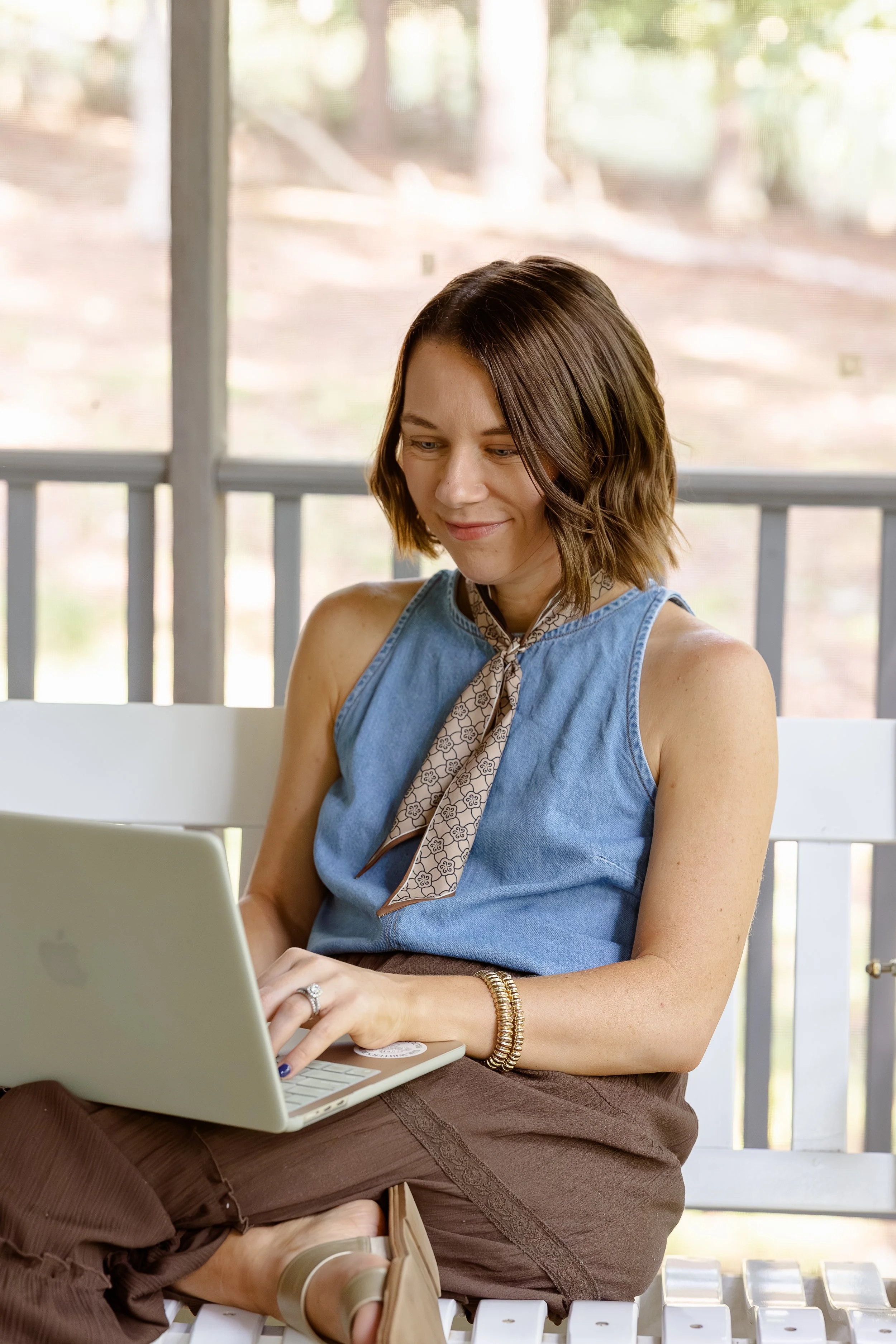 Natty Writes, blog writing strategist, sitting outside on a white chair in the daytime, smiling at a laptop on her lap, in front of a screened porch with a blurred view of trees and greenery in the background.