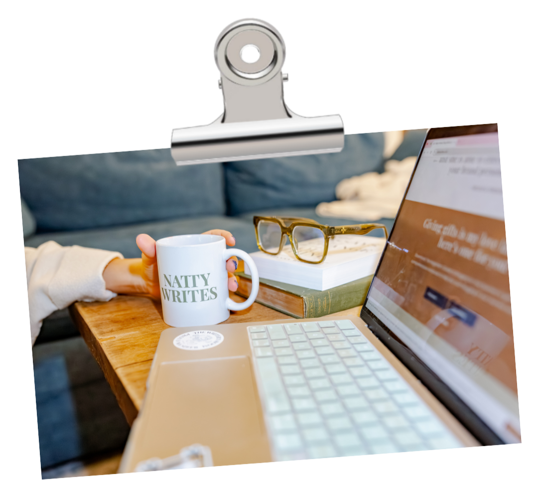 Close-up of a wooden desk with Natty, blog writer, holding a white mug that says 'NATTY WRITES', a stack of books, a pair of glasses, and an open laptop displaying a website, in a cozy room setting.