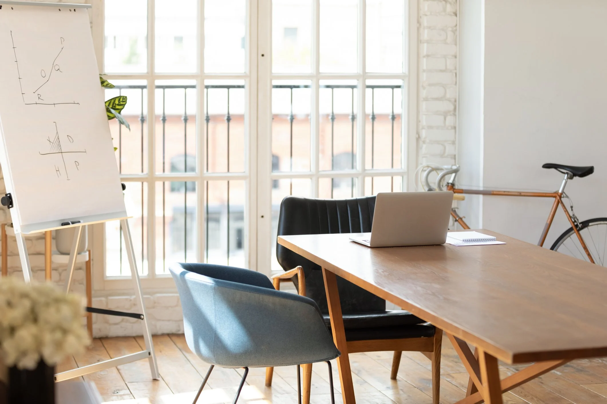 Bright office with large windows, a wooden table with a laptop and notebook, blue and black chairs, a whiteboard with graphs, a bicycle against the wall, and potted plants.