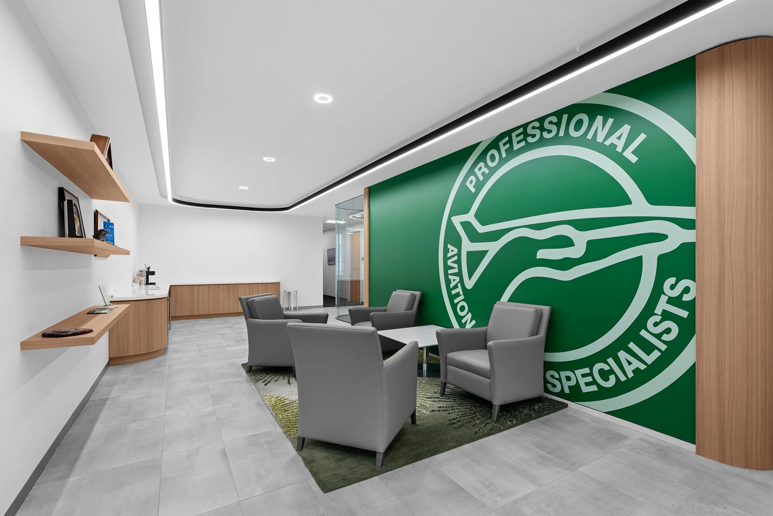 Modern office lounge with four gray armchairs and a small white table on a green rug, large green wall with an airplane logo and the words "Professional Aviation Specialists," wooden accents, and white walls with floating wooden shelves.