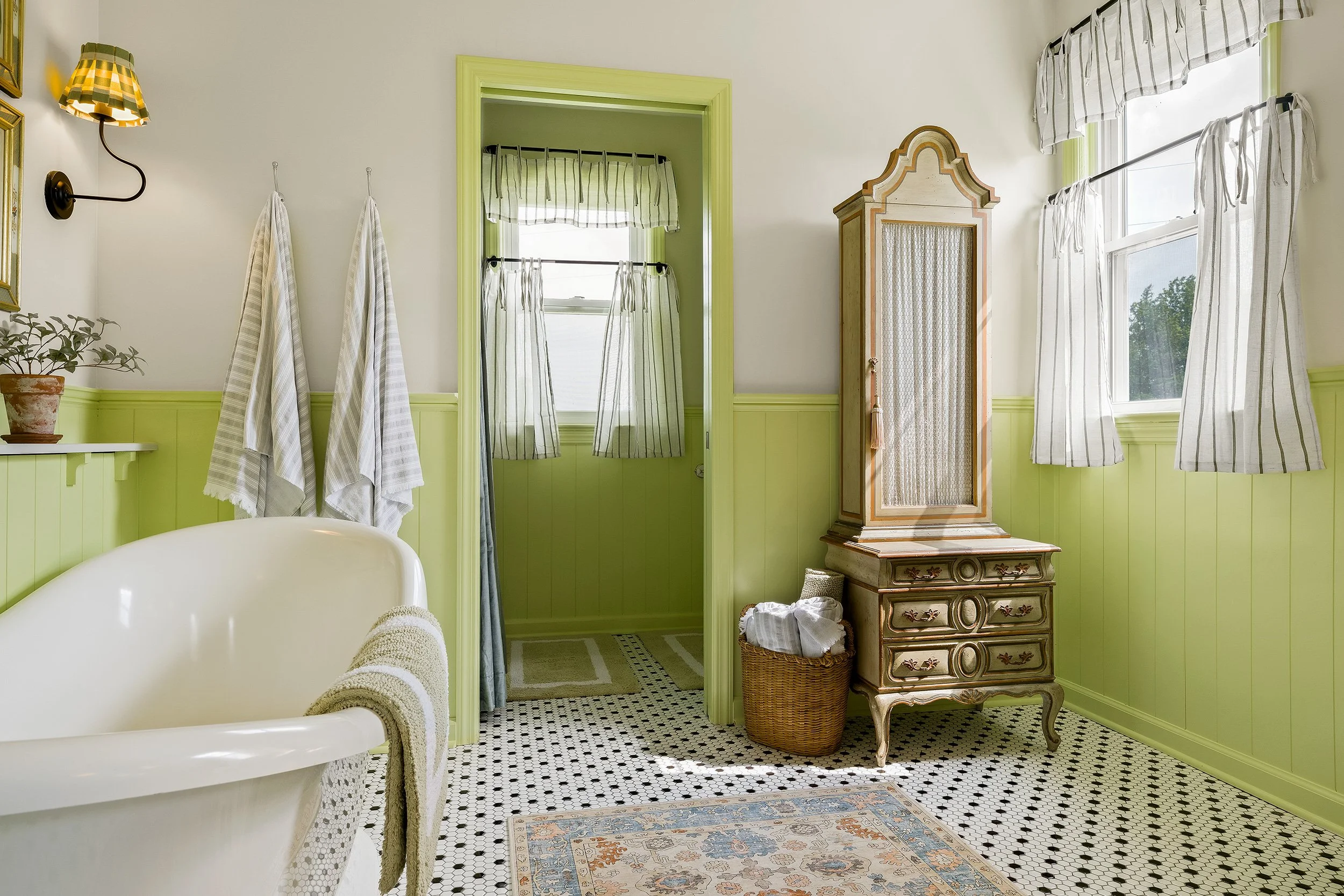 Bright vintage bathroom with a clawfoot tub, green wainscoting, black and white tiled floor, wooden chest, windows with striped curtains, and towels hanging on hooks.