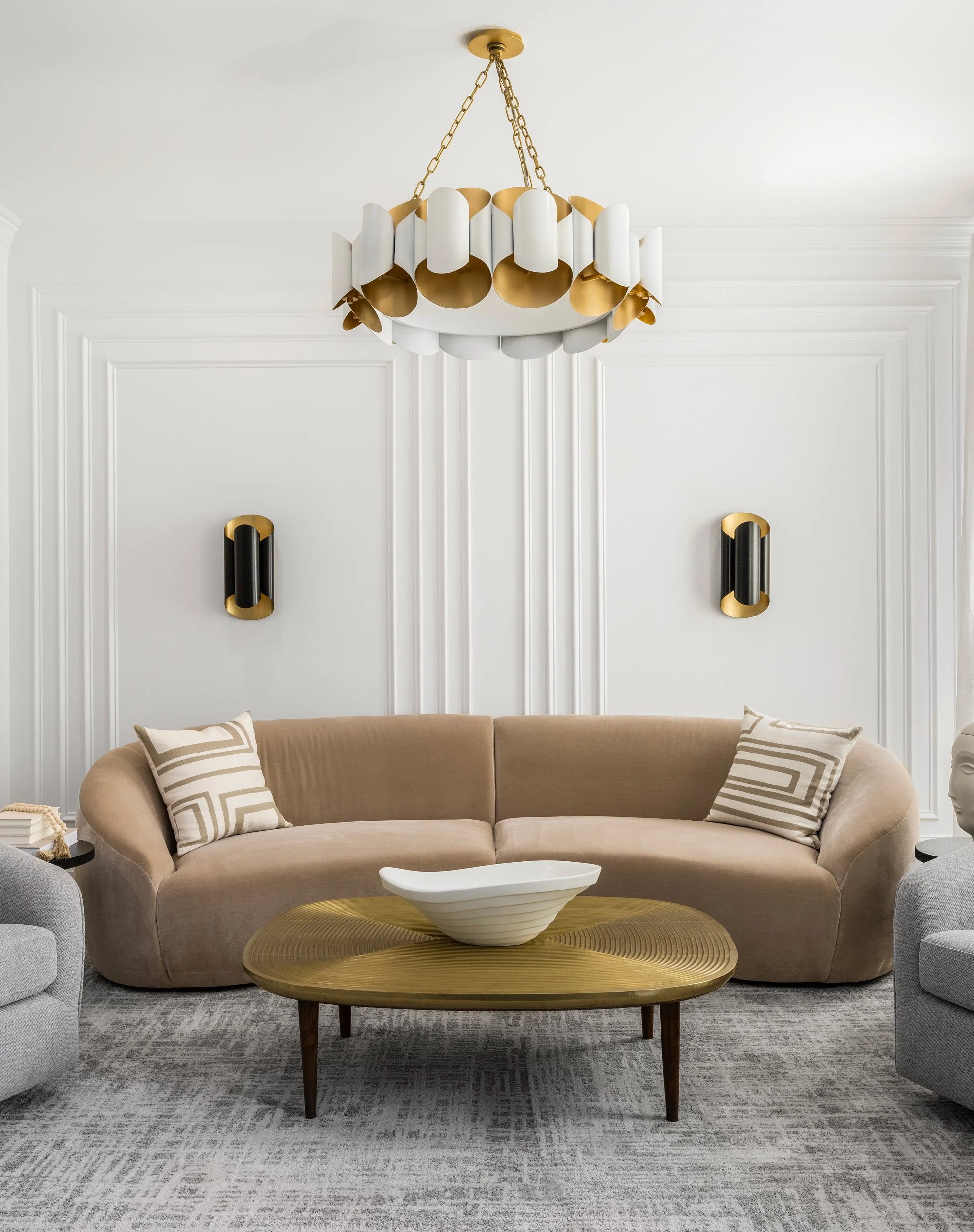 Elegant living room with a beige velvet sofa, decorative pillows, a wooden coffee table with a white ceramic bowl, wall sconces, and a statement chandelier.