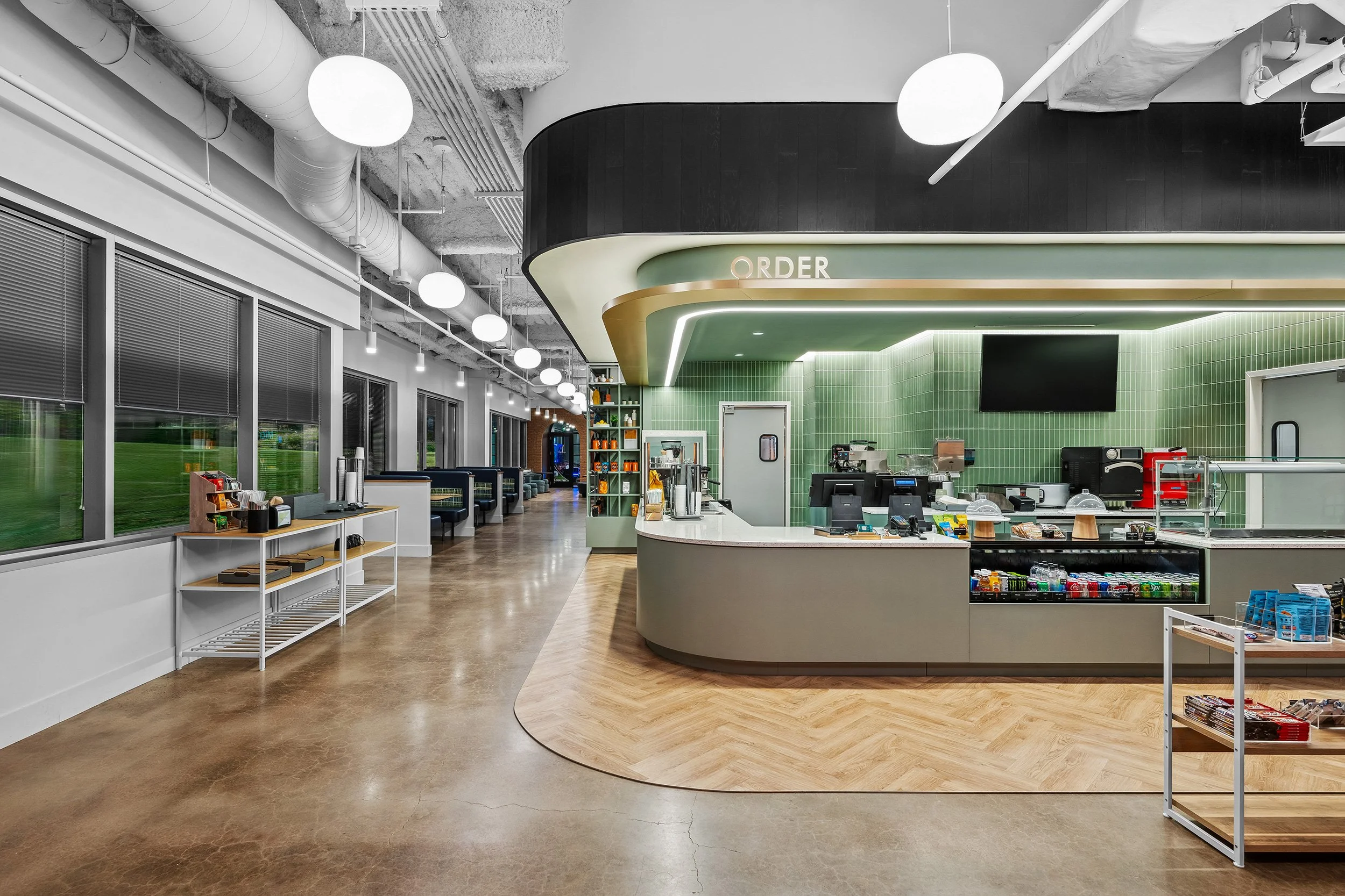 Interior of a modern convenience store or café with a curved counter, green tiled walls, and a seating area with booths and large windows.