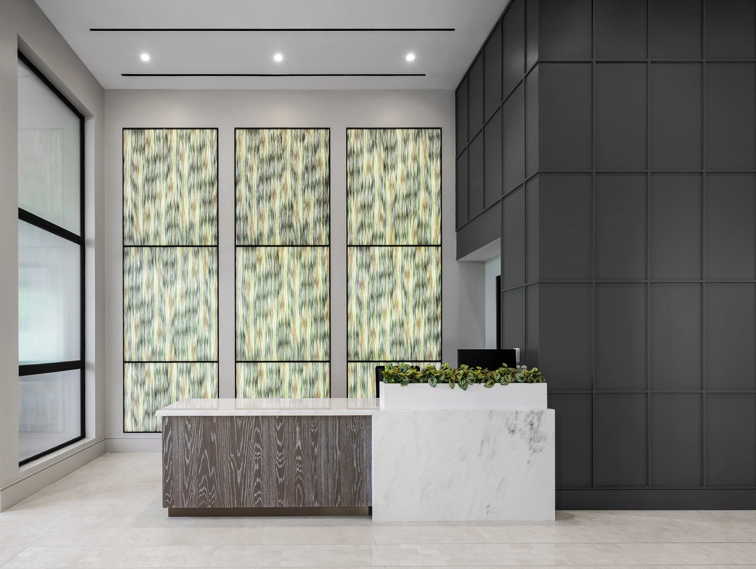 Modern lobby reception area with a marble desk, black computer monitor, decorative plant, large stained glass windows, and a dark gray wall with a geometric pattern.