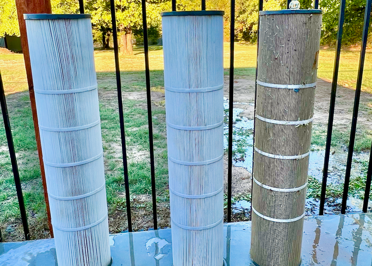 Three pool filter cartridges, two clean and one dirty, standing vertically on a wet surface with a metal fence and grass in the background.