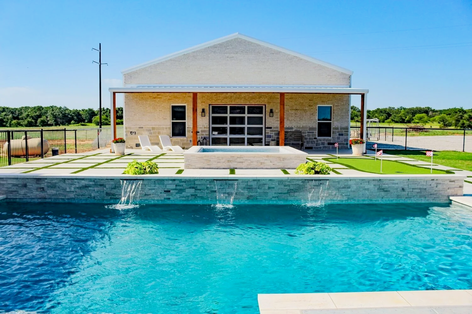 Backyard with swimming pool, patio, lounge chairs, and a house with a shaded porch, set in a rural landscape. Custom backyard pool built by Barefoot Pools, serving Bridgeport, Decatur, Weatherford, and Springtown, Texas.