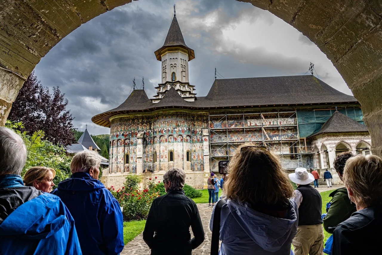 Tourists gathered around in front of a church or monastery with painted exterior walls, scaffolding, and a tall spire under a cloudy sky.