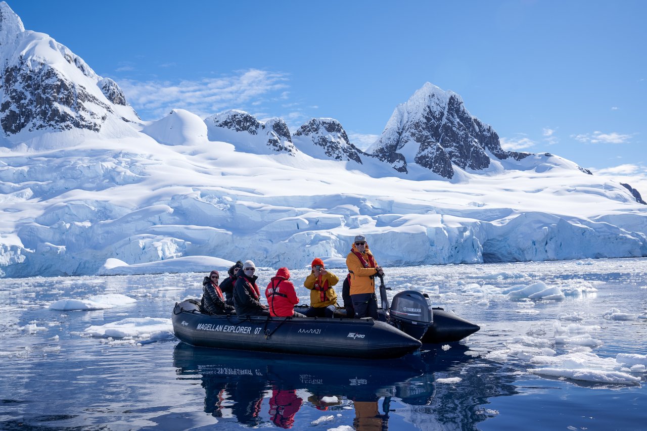 A group of six people on a black inflatable boat in icy water with snow-covered mountains and glaciers in the background.
