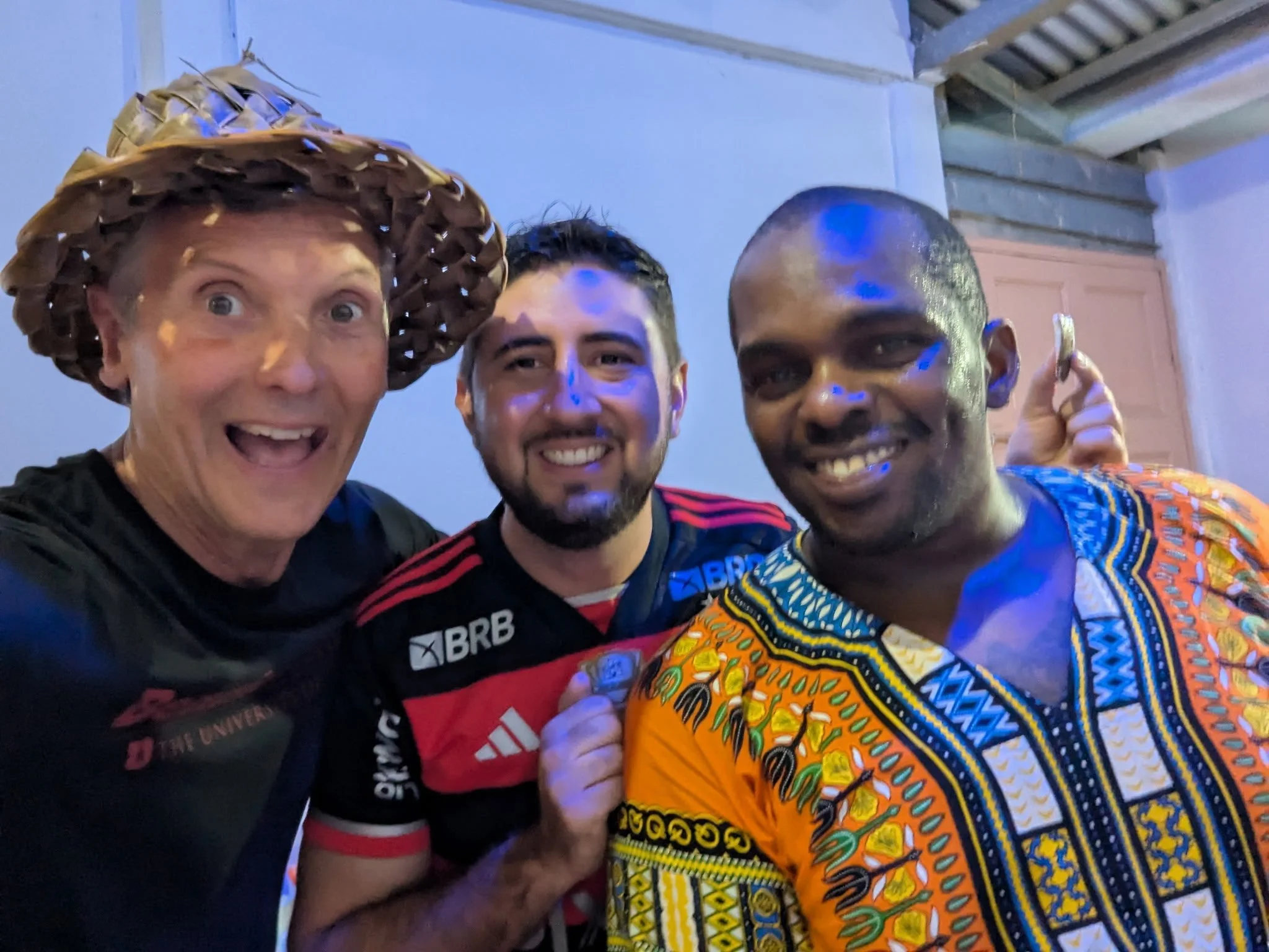 Three men smiling, taking a selfie together. The man on the left is wearing a hat made of woven material. The man in the middle is wearing a sports jersey with sponsor logos. The man on the right is wearing brightly colored traditional African clothing. They are indoors with a background showing a ceiling with exposed beams.