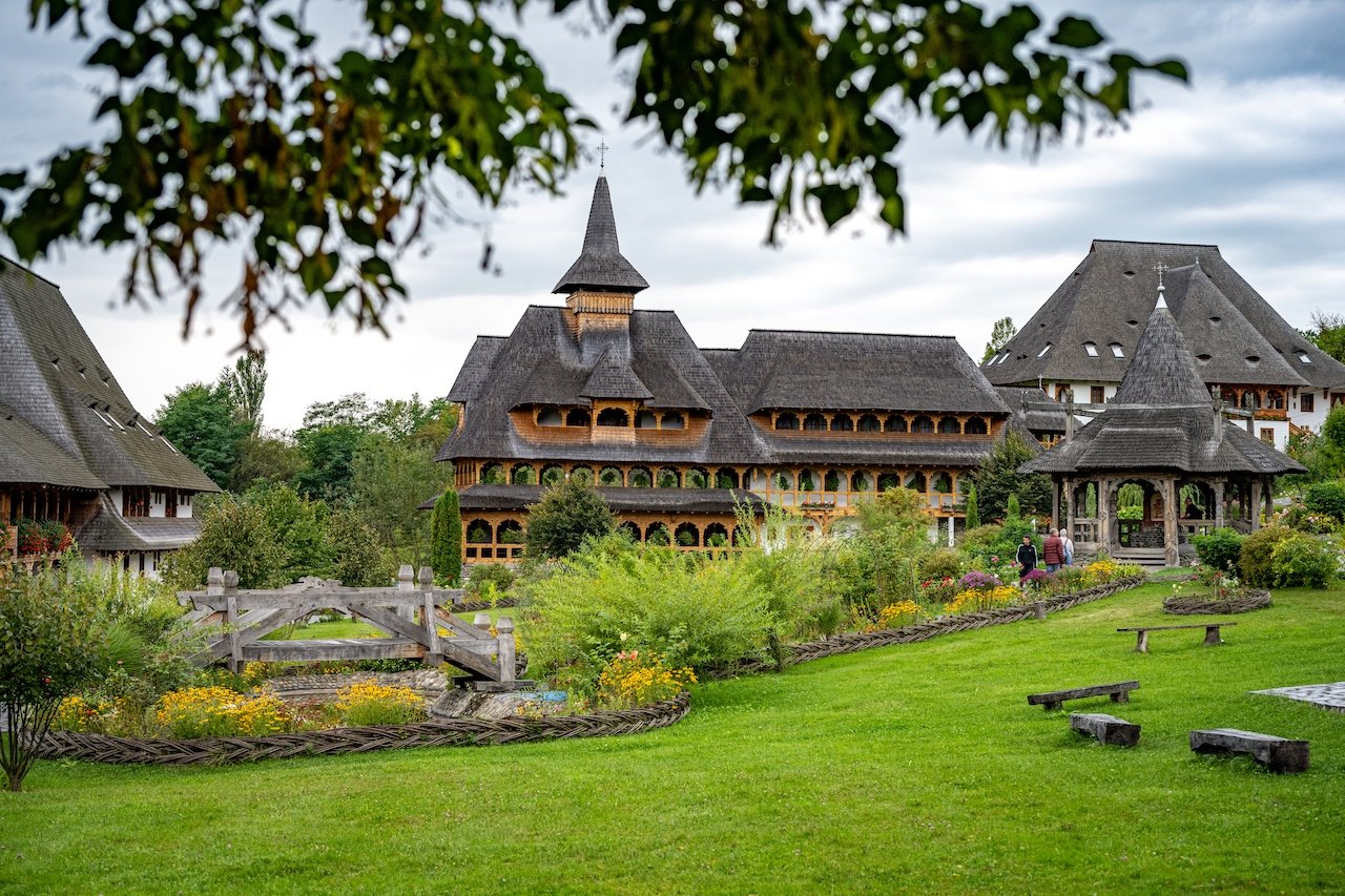 A large traditional wooden building with multiple peaked roofs, surrounded by a lush garden with colorful flowers, trees, and walking paths.