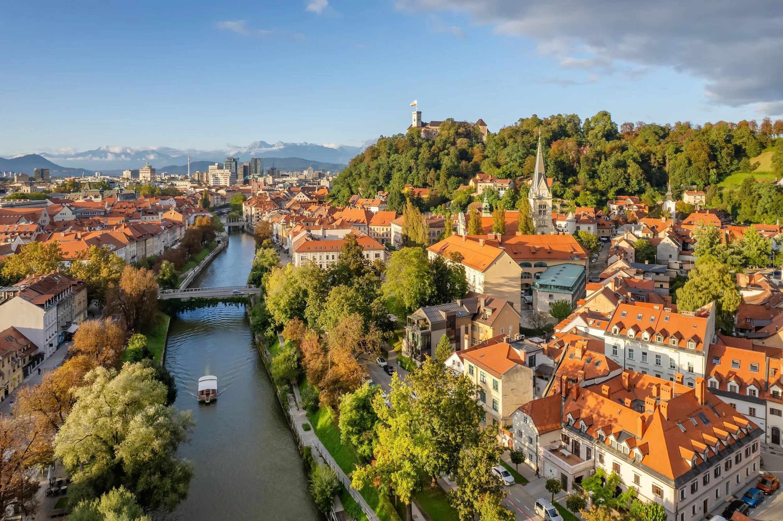 Aerial view of a European city with orange rooftops, a river with a boat, and a hill with a castle and church spire.