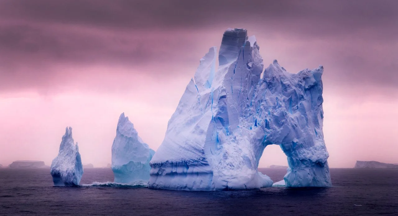 Large iceberg in the ocean with a natural arch formation, against a pink and purple cloudy sky.