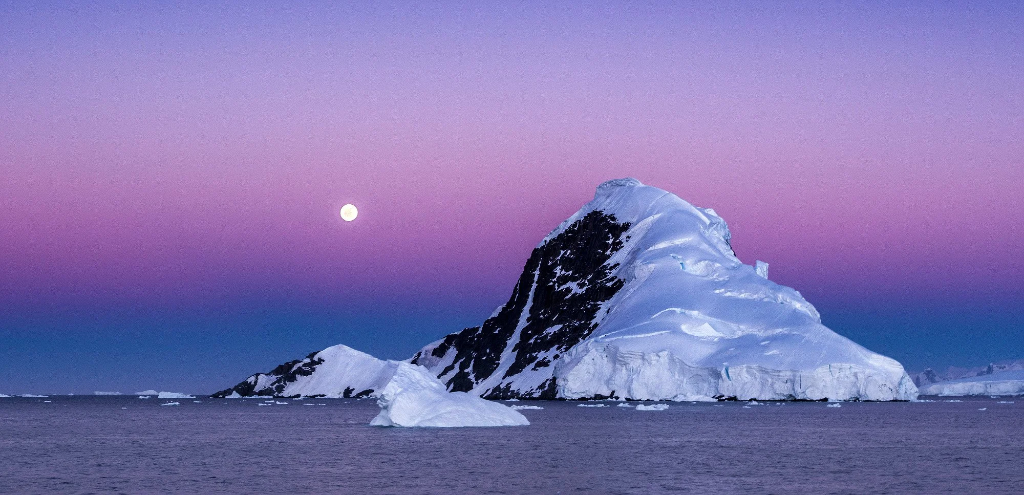 A snow-covered mountain in Antarctica with a large ice formation in the ocean in front and a full moon in the sky with purple and blue hues.