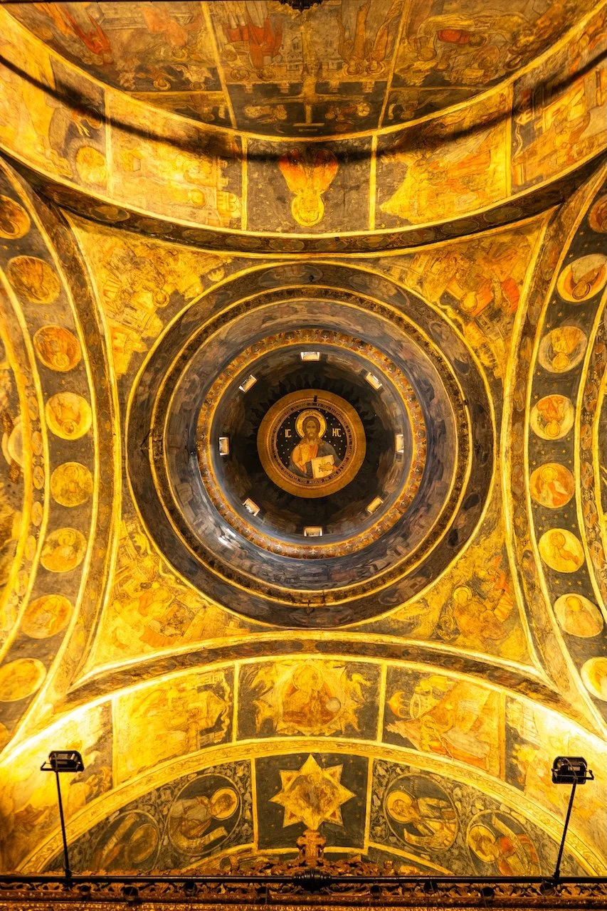 Looking up at a domed ceiling painted with religious icons and artwork in golds and dark hues, featuring a central image of Christ holding a book surrounded by decorative patterns.