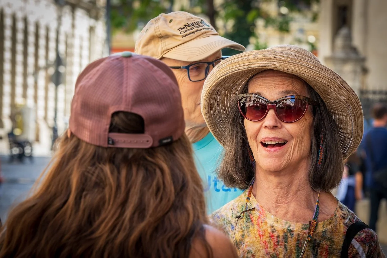 Three women talking outdoors; one is wearing a wide-brimmed hat and sunglasses, another has a baseball cap, and the third has a beige hat with a logo, with blurred urban background.