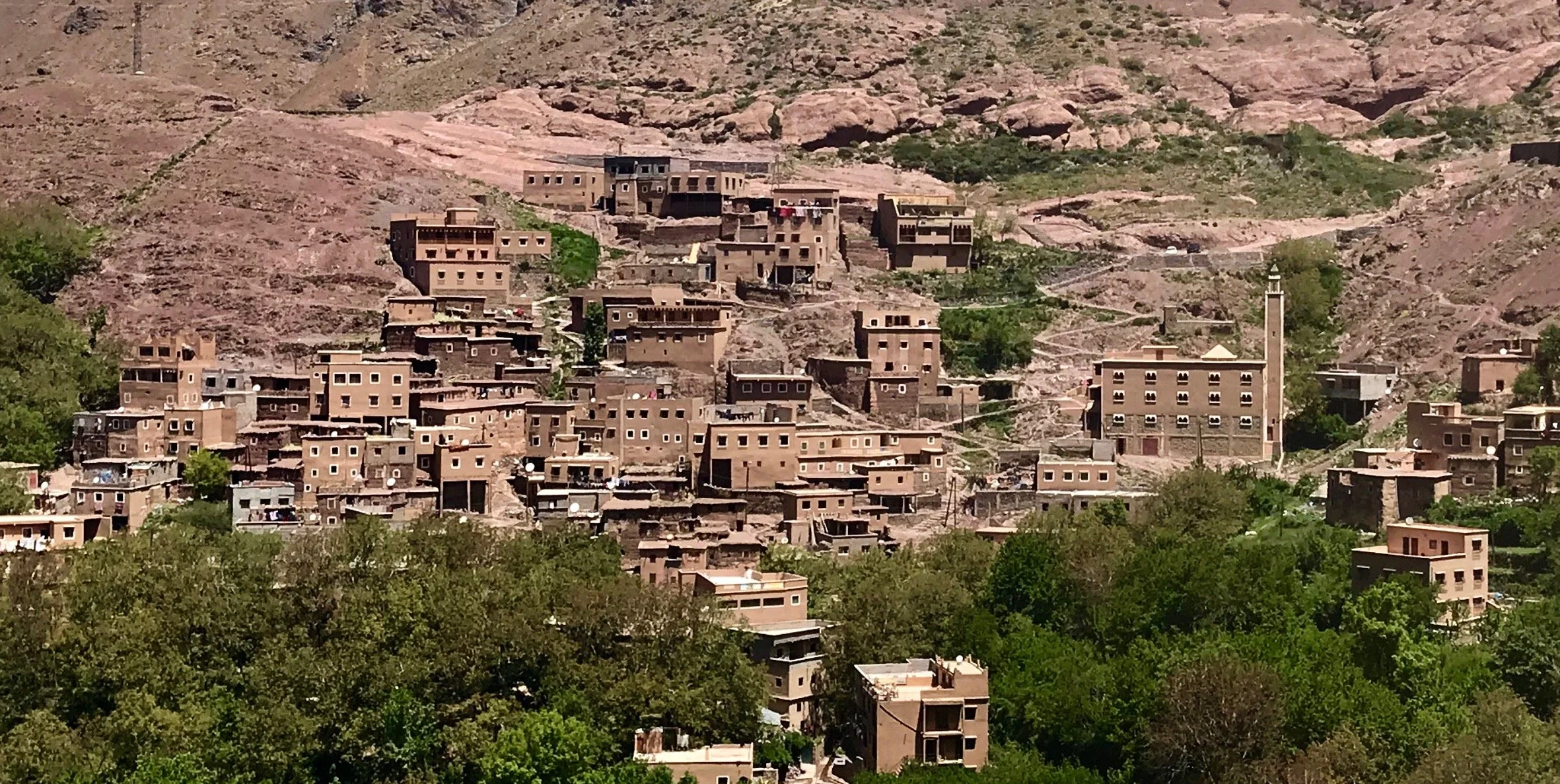 Hillside village with numerous tan-colored buildings, some with flat roofs, surrounded by greenery and a few trees, with a mosque featuring a tall minaret, set against arid, rocky terrain.