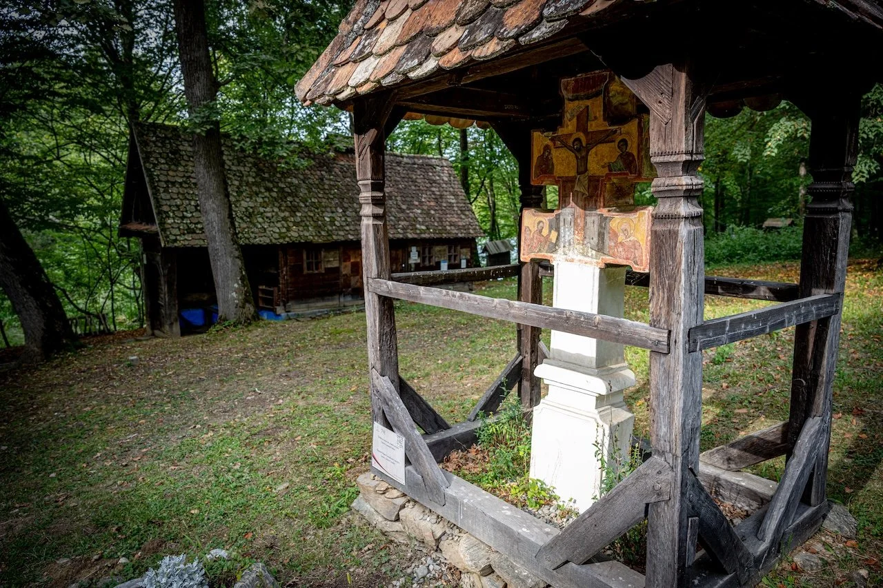 A small wooden outdoor shrine with religious icons, vandalized with a broken cross, surrounded by a wooden fence, in a wooded area with two small wooden cottages in the background.