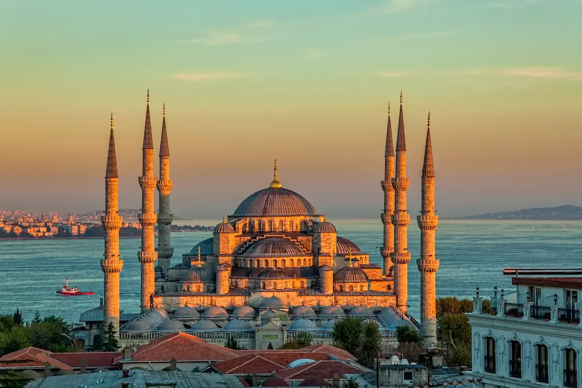 An evening view of the Sultan Ahmed Mosque (Blue Mosque) in Istanbul, Turkey, with its multiple minarets and large central dome, set against the Bosphorus Strait and a distant cityscape.