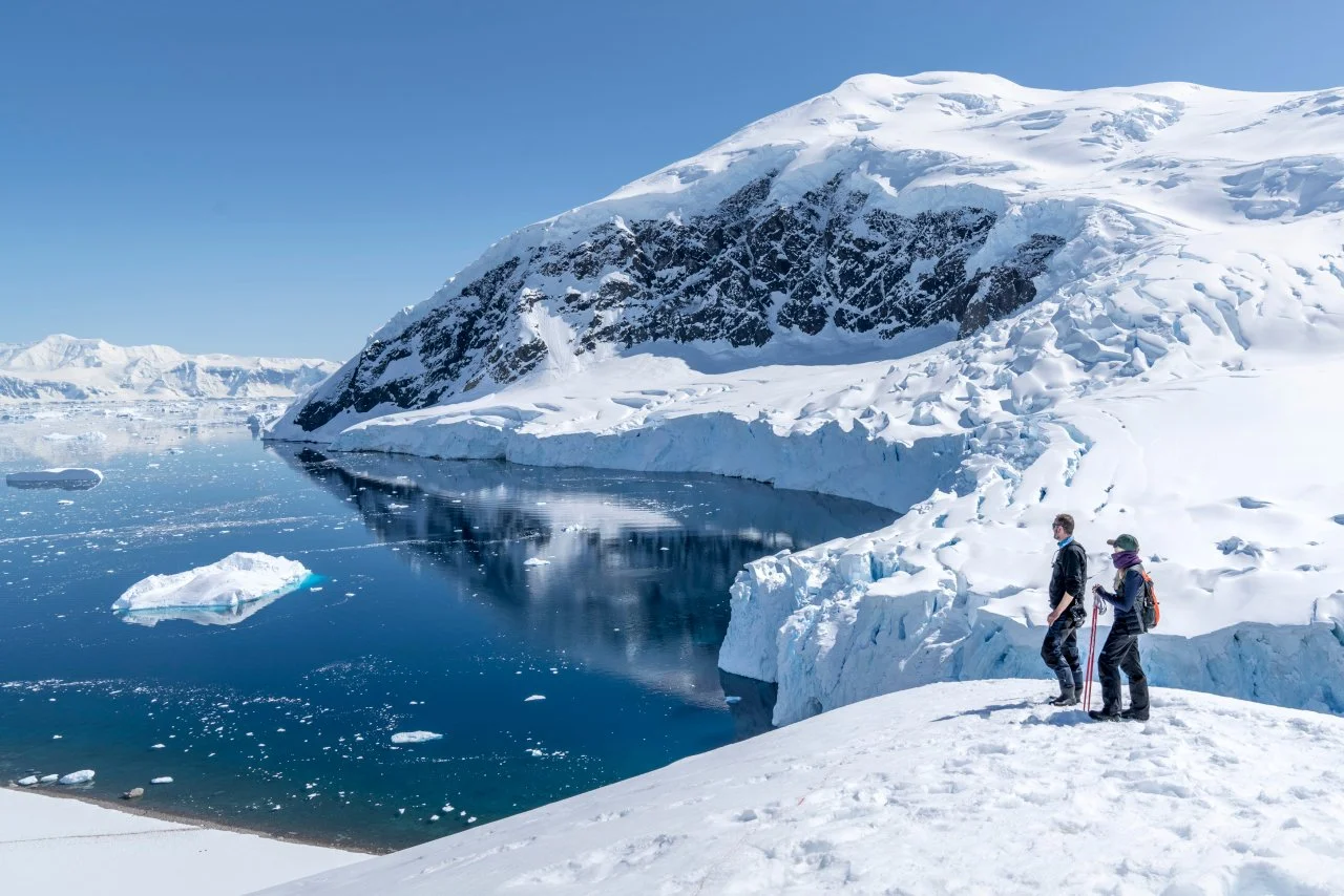 Two people in winter clothing hiking on snowy terrain near a glacier with icy waters and floating icebergs under a clear blue sky.