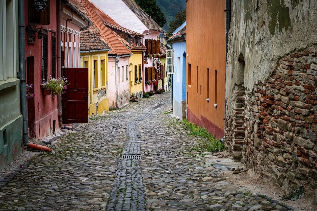 A narrow cobblestone street in a colorful European village, lined with pastel-colored houses with flower boxes, shutters, and open doors.