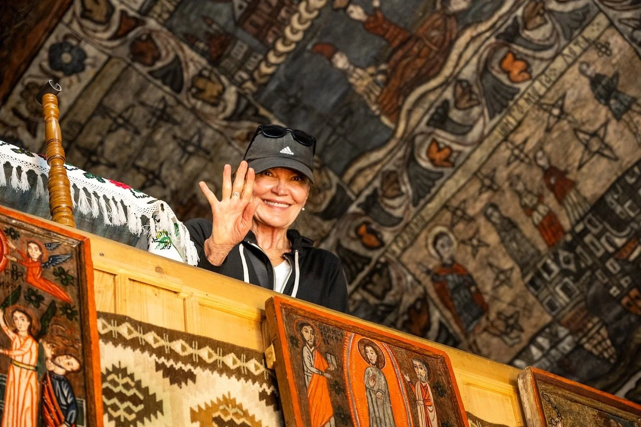 An elderly woman wearing a black hat and black jacket waves from a wooden balcony, surrounded by traditional religious and cultural artwork and textiles inside a decorated space with a detailed, ornate ceiling.