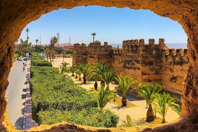 View of an ancient stone fortress wall with palm trees and a sidewalk, seen through a rough stone window opening, in a sunny desert setting.