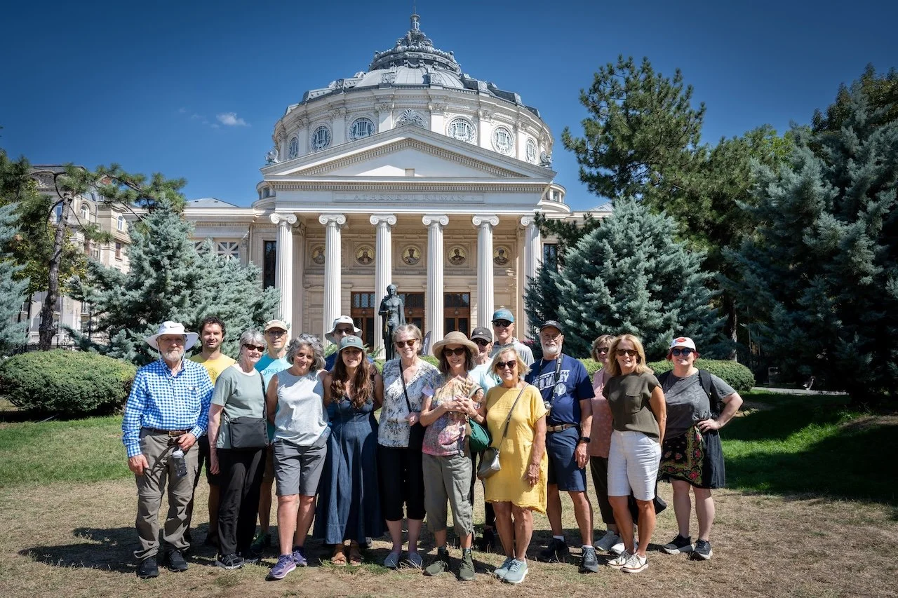 A group of people posing outdoors in front of a historic building with columns and a domed roof, surrounded by trees, on a sunny day.