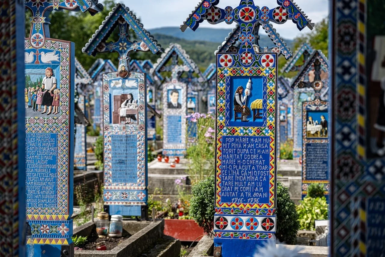 Colorful, decorated grave markers with religious and patriotic artwork, located in a cemetery. The markers feature images of Jesus Christ, a woman praying, and text in Romanian, surrounded by greenery.