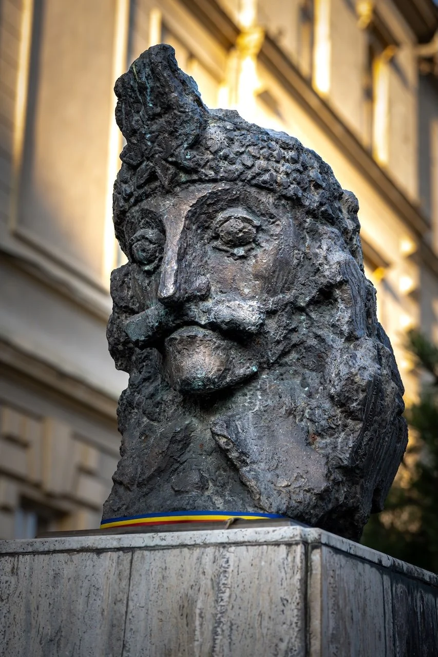 Bronze bust sculpture of a historical figure with a prominent mustache and mustache, mounted on a stone pedestal with a Romanian flag ribbon, situated outdoors with a building in the background.