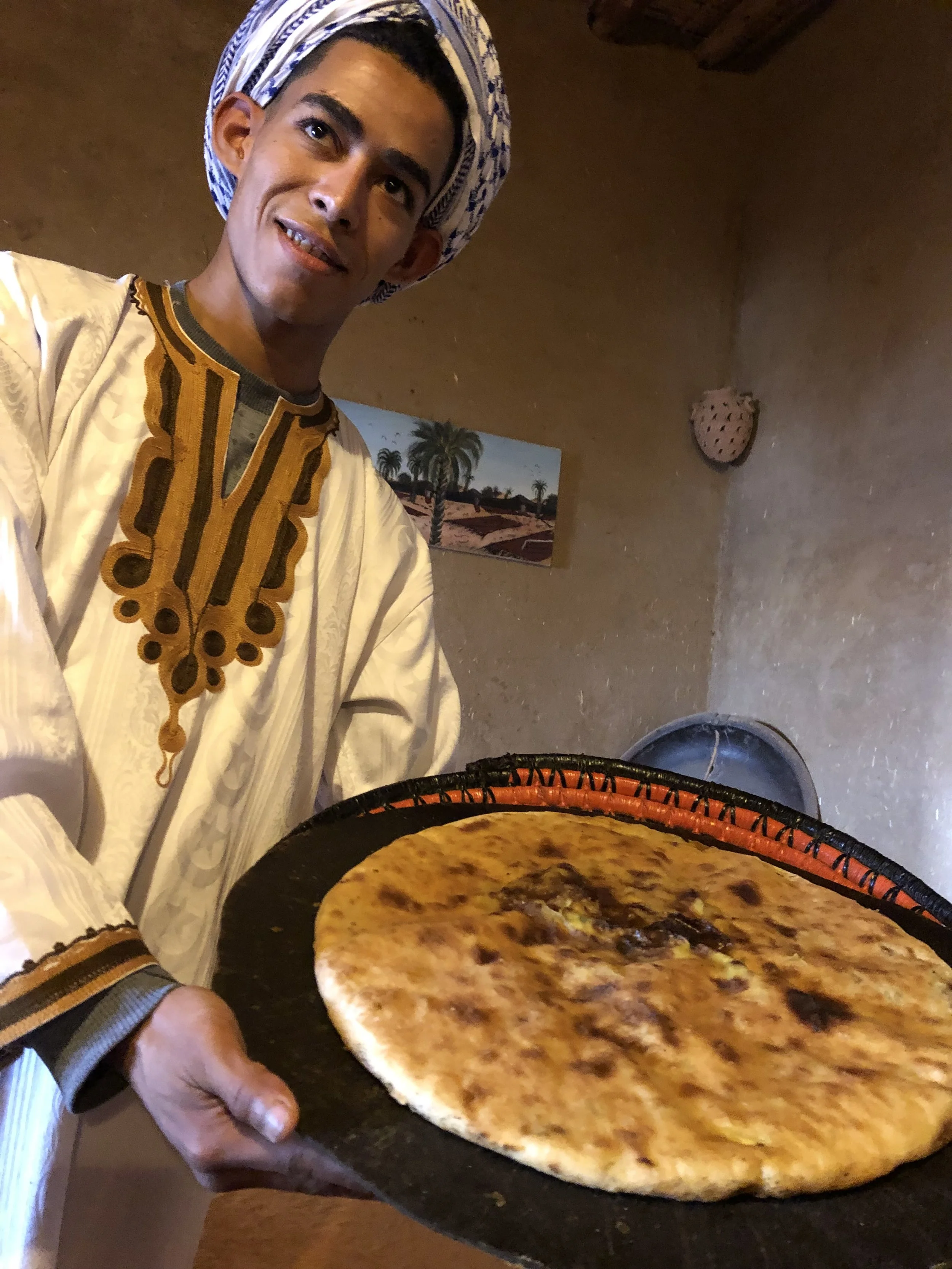 A man wearing traditional Moroccan attire and a headscarf holding a large pizza on a black serving tray inside a room.