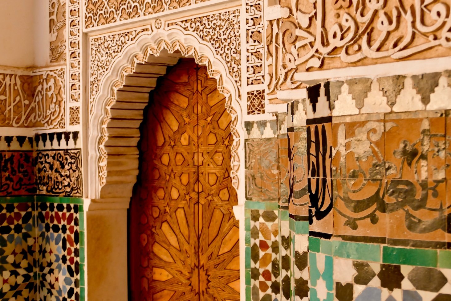 Decorative wooden door with intricate geometric carvings, surrounded by colorful Islamic tile patterns and Arabic calligraphy.
