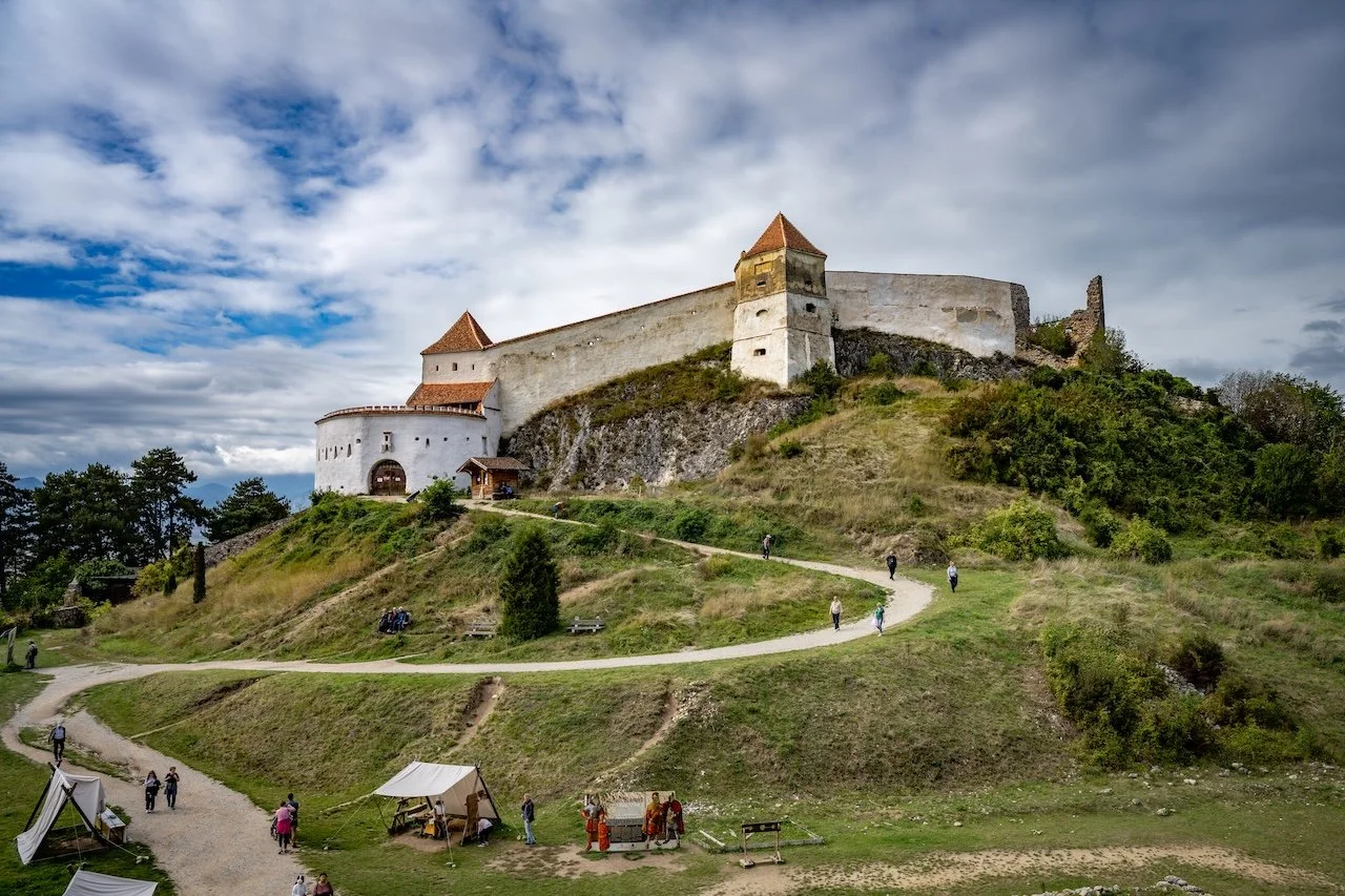 A medieval castle with white walls and red-tiled roofs sits atop a grassy hill, with a winding path leading up to it. There are people walking around the grassy area near the castle and a few tents set up at the base.