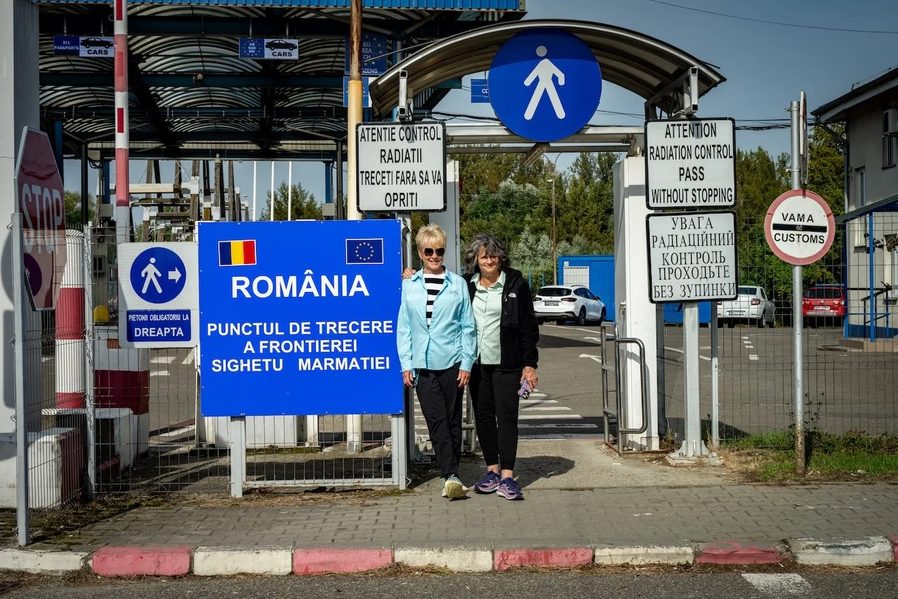 Two women standing in front of the Romanian border crossing sign with a blue background and the Romanian flag, near a pedestrian walkway. There are multiple signs indicating border control procedures, a stop sign, and a customs sign. Several cars are parked behind the gate.