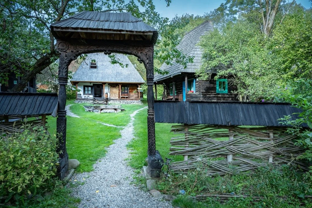 A village scene with traditional wooden houses, a decorative archway made of carved wood, and a gravel pathway leading to the houses, surrounded by lush greenery.