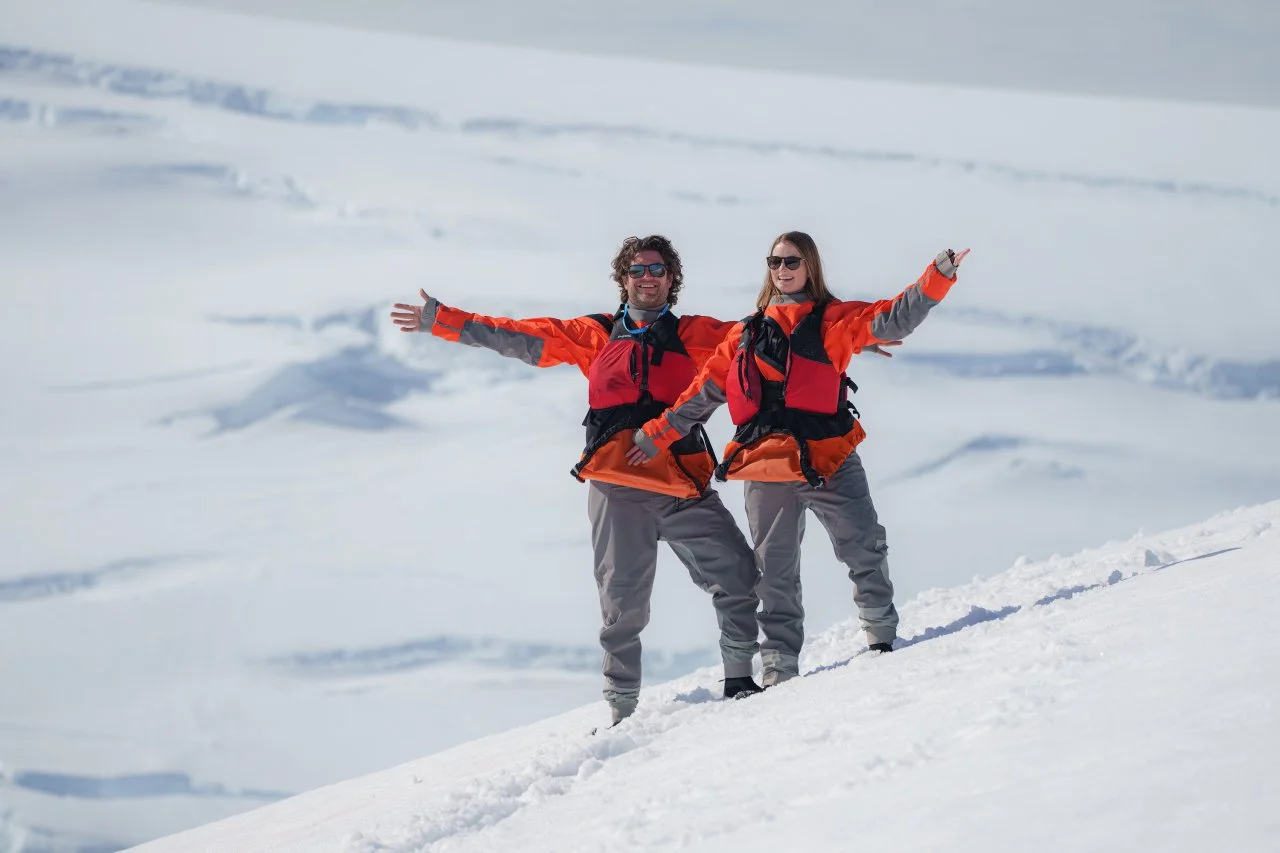 Two people in orange and gray winter jackets, gray pants, and sunglasses standing on snow-covered ground with arms outstretched, smiling, in a snowy landscape.