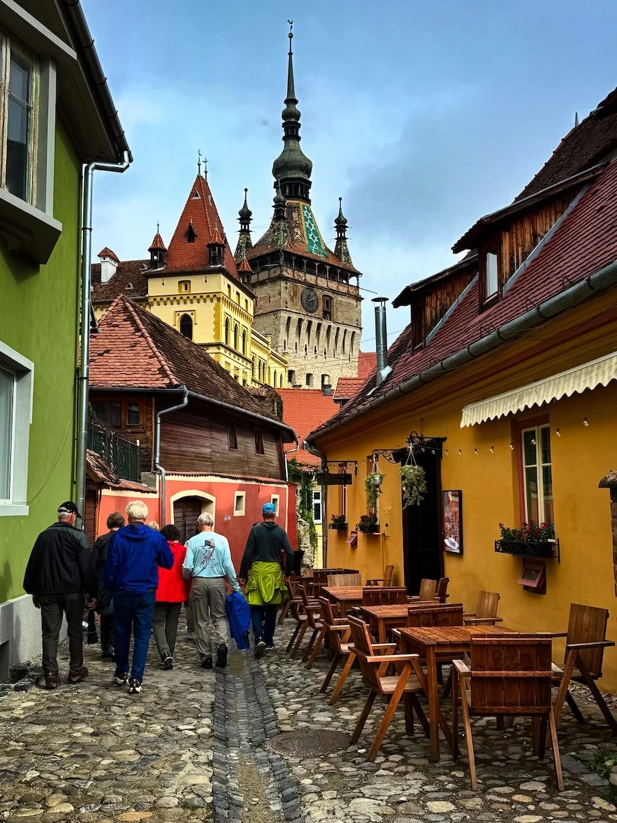A cobblestone alleyway with colorful buildings on each side, outdoor tables and chairs, and a group of people walking towards a historic clock tower with a tall spire in the background.