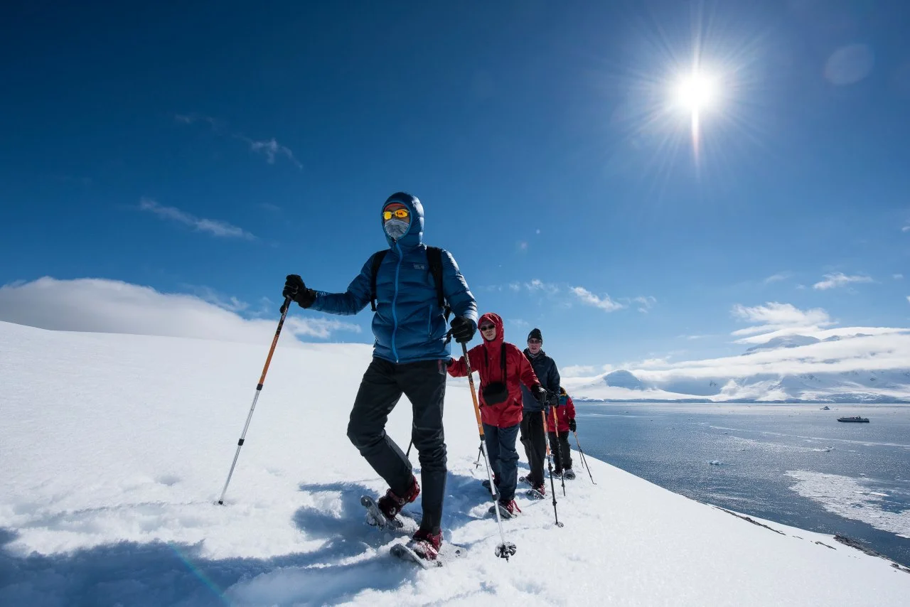 A group of four people wearing winter gear and snowshoes, hiking on a snowy landscape near water under a bright sun with a clear blue sky.
