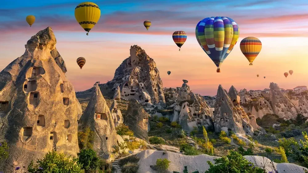 Hot air balloons flying over rocky formations and unique cave dwellings during sunrise or sunset.
