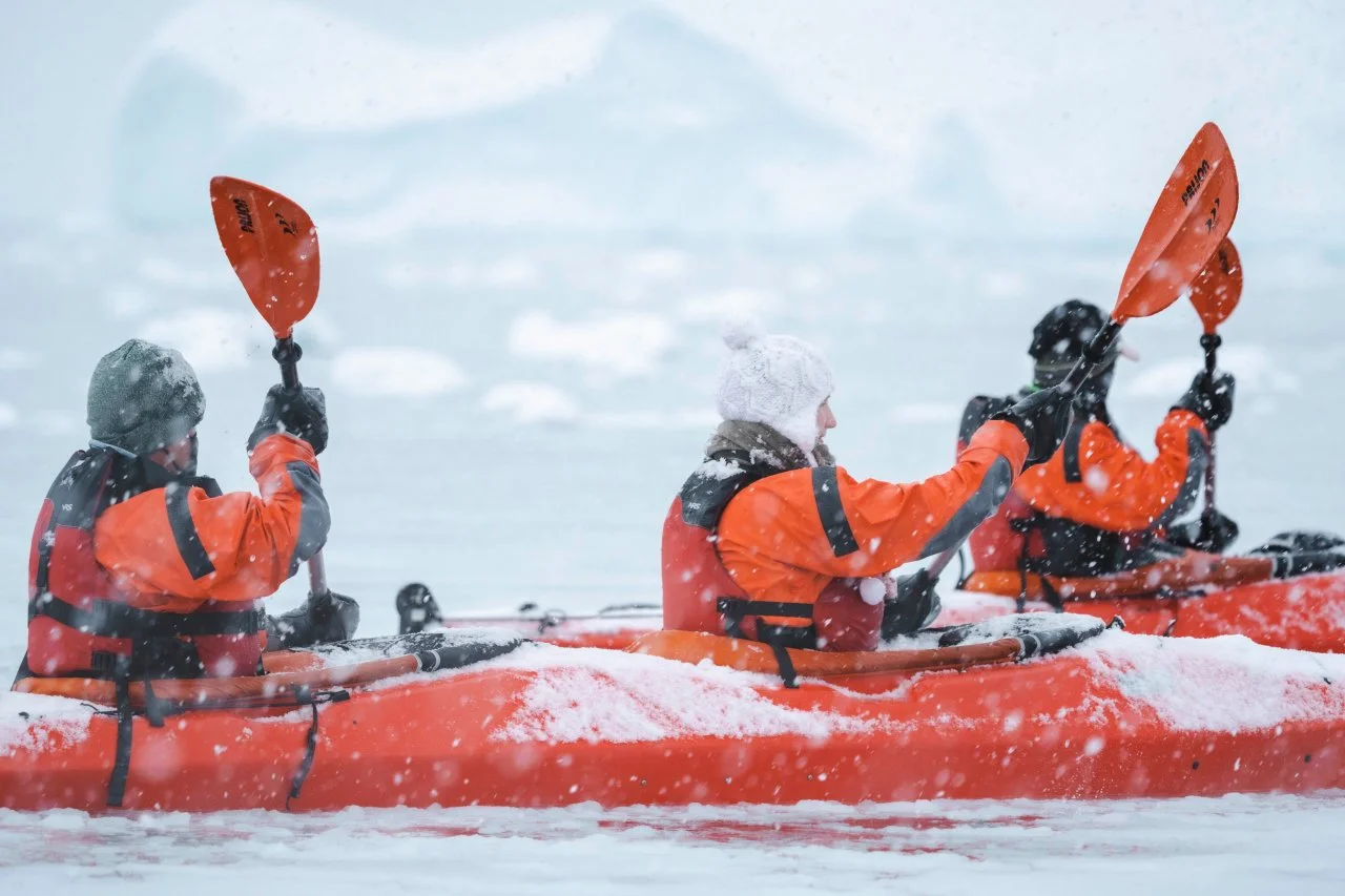Three people in orange and black jackets kayaking through icy waters in snowy weather, with snow on their kayak and paddles.
