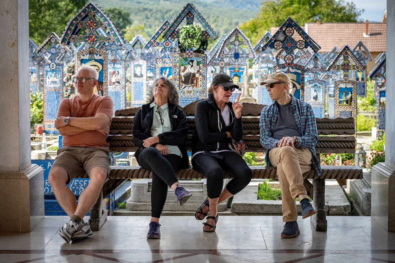 Four tourists sitting on a wooden bench inside a church, with colorful mosaic grave markers in the background.