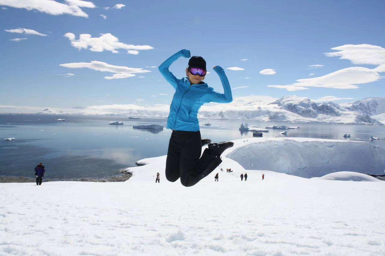 Person jumping in snowy landscape with mountains and icebergs in the background, wearing a blue jacket and ski gear.