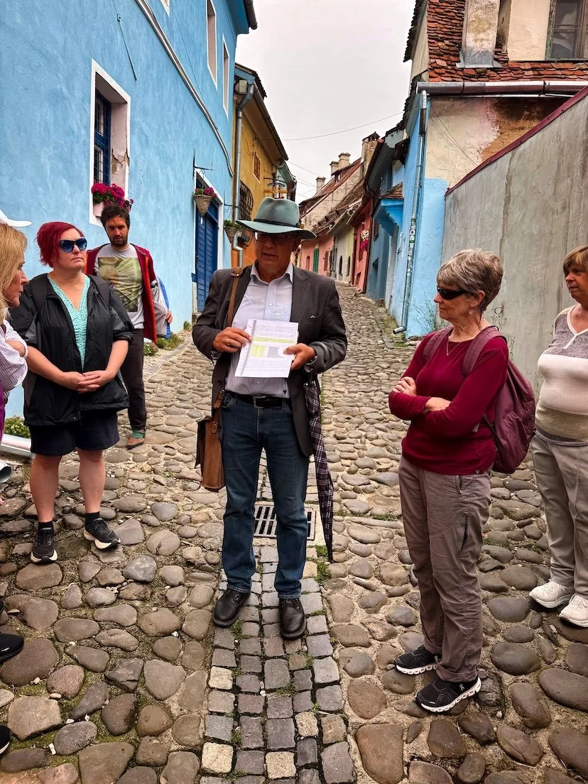 Group of people listening to a man in a suit and hat holding papers on a cobblestone street with colorful buildings.