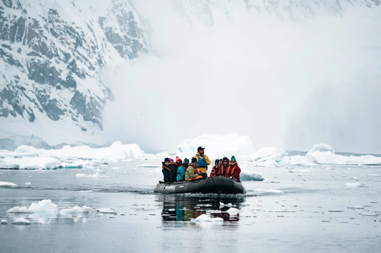 Group of people in a black inflatable boat navigating icy waters with icebergs and snow-covered mountains in the background.