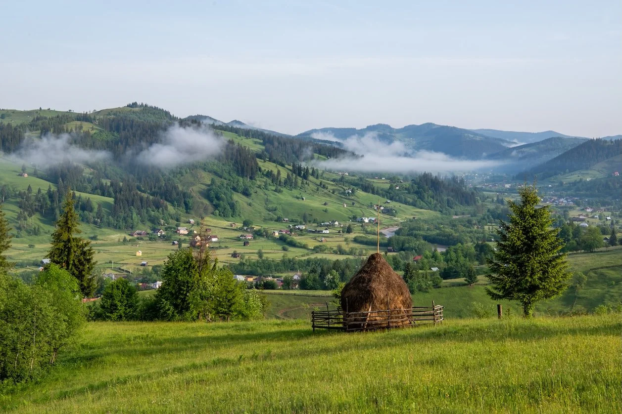 A rural landscape with green rolling hills, scattered houses, trees, and fog in the background, with a haystack and wooden fence in the foreground.