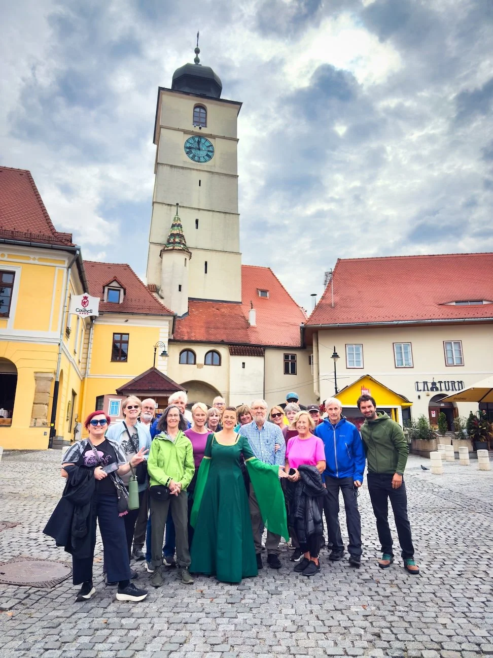 Group of people standing on cobblestone street with historic clock tower and colorful buildings in the background during daytime.
