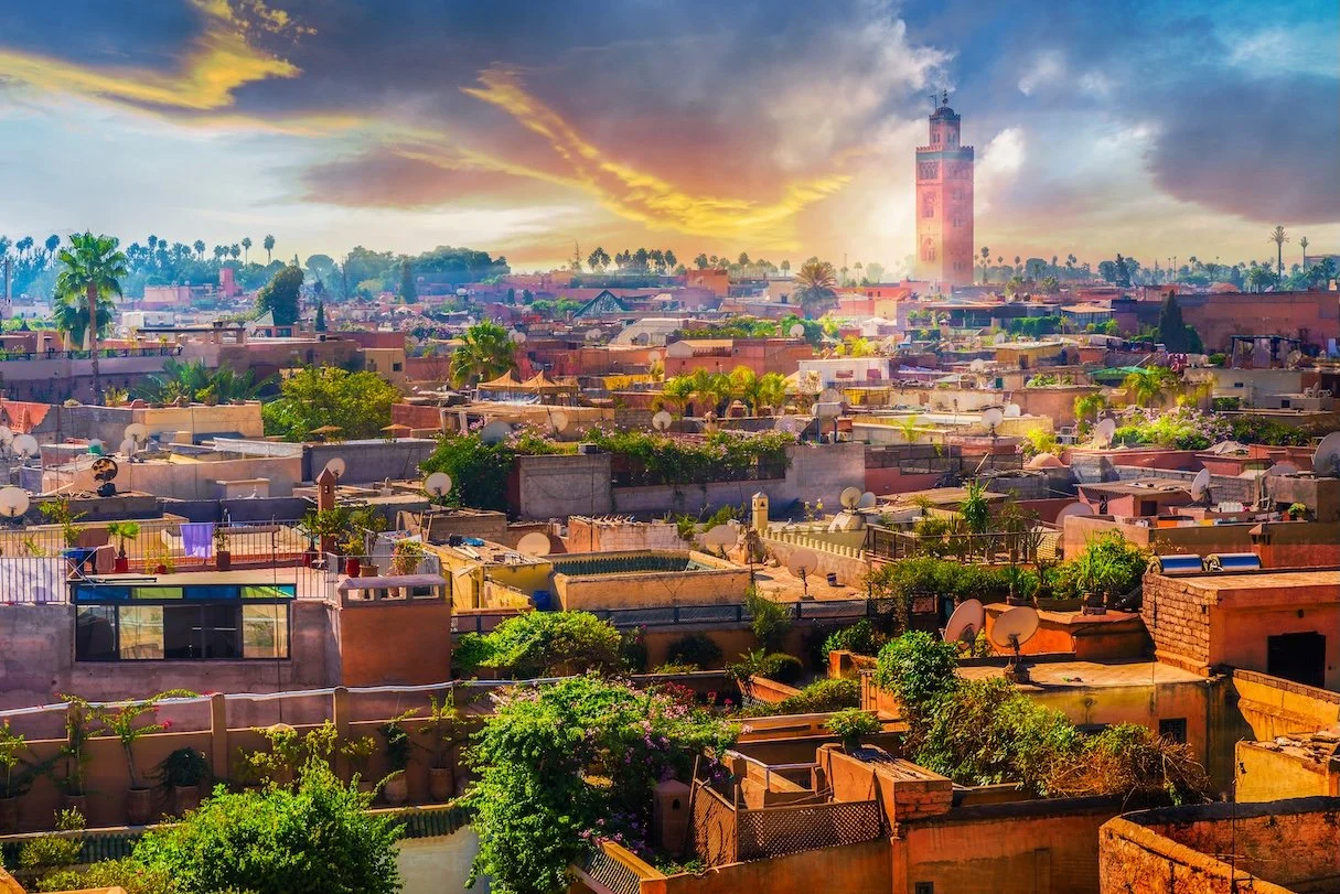 Cityscape of Marrakech, Morocco at sunset with the Koutoubia Mosque's minaret in the background, surrounded by buildings, satellite dishes, and greenery.