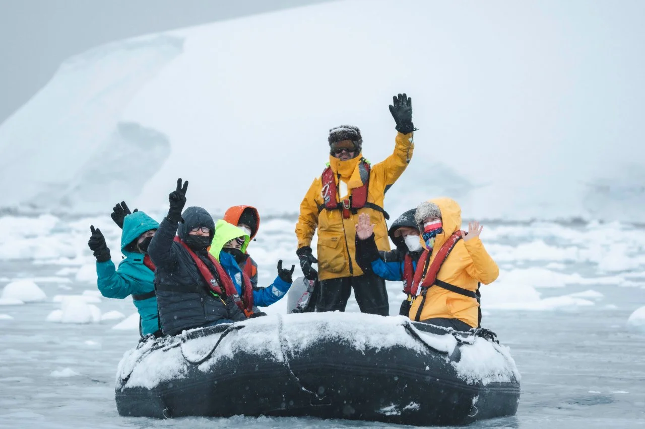 A group of six people wearing winter gear and life jackets on an inflatable boat navigating icy waters, with snow and icebergs in the background.