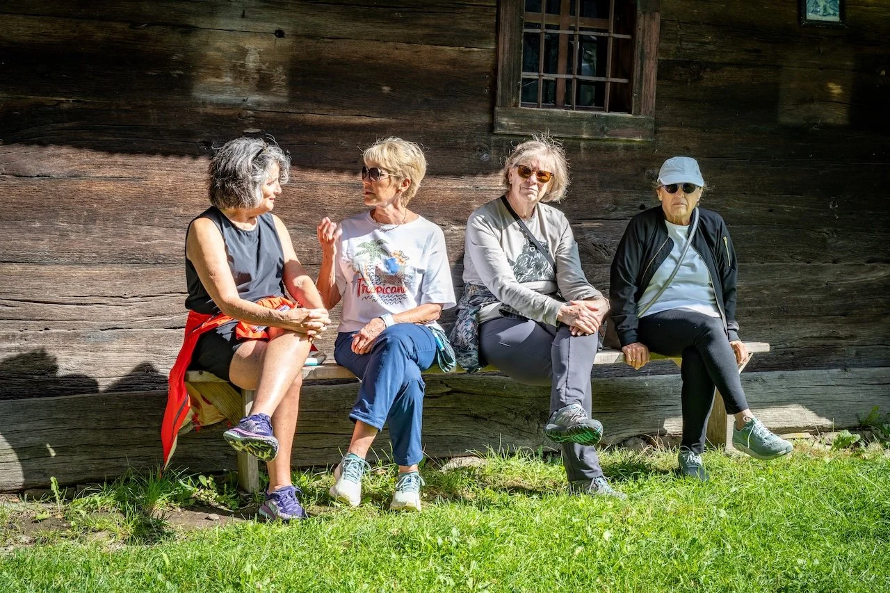 Four women sitting on a wooden bench outside a rustic wooden building, engaged in conversation.