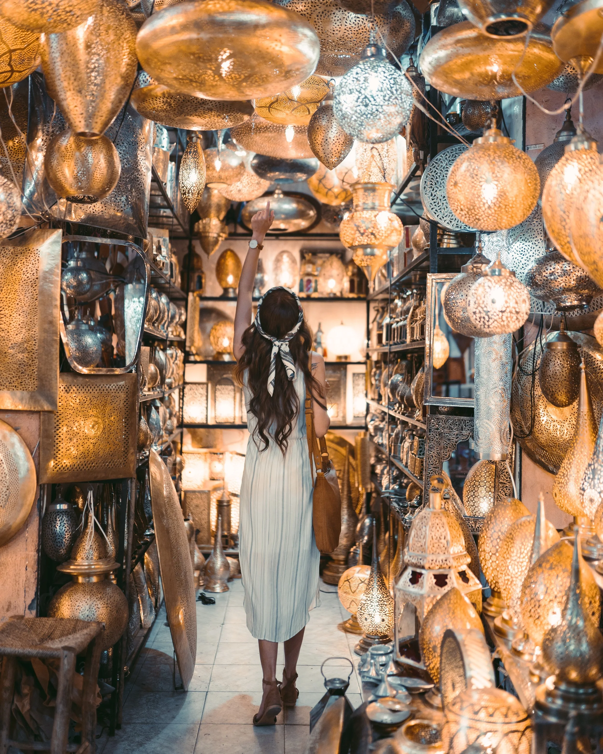 A woman shopping in a store filled with decorative lamps and lanterns, reaching up to examine them.